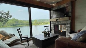 A cozy living room with a stone fireplace, a large window overlooking a lake, and comfortable seating. - The Water’s Edge * * Unique Muskoka Treehouse * * (Utterson)