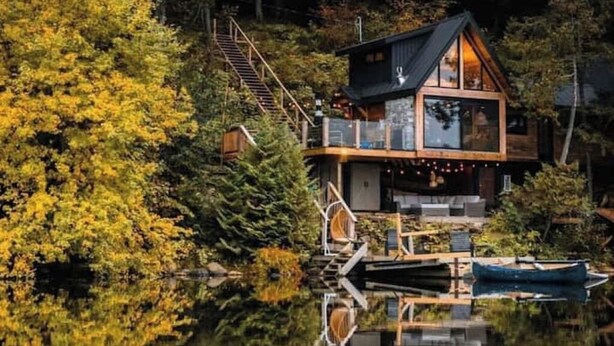 A cabin with a glass facade, wooden deck, and a dock extending into a lake surrounded by trees. - The Water’s Edge * * Unique Muskoka Treehouse * * (Utterson)