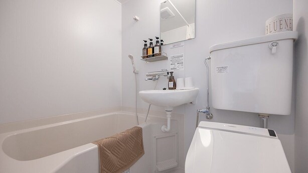A modern bathroom with a bathtub, a sink, and a toilet. There are toiletries on a shelf and a towel hanging on the wall. - HOTEL R9 The Yard Honjowakaizumi (Honjo)