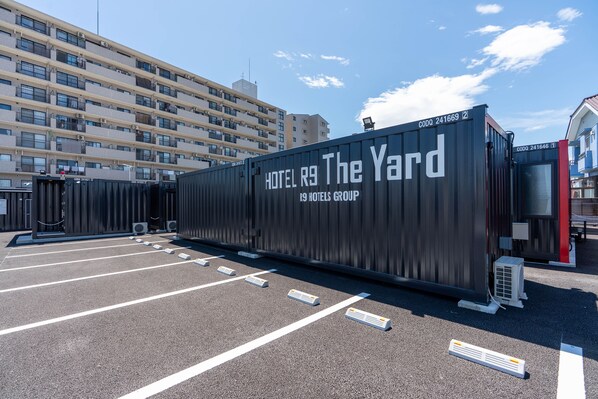 Un hotel moderno tipo contenedor, grande, con el texto "HOTEL R9 The Yard" y "R9 HOTELS GROUP" en su lateral, estacionado en un estacionamiento con señalización clara.