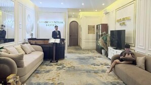 A hotel lobby with a reception desk, sofas, a television, and a decorative wall. - Grand Central Hotel- Vinhomes Ocean Park 3 (Nghia Tru)