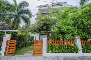 Un edificio de varios pisos con fachada blanca, entrada protegida por puertas de madera y exuberante vegetación, incluyendo palmeras y plantas con flores.