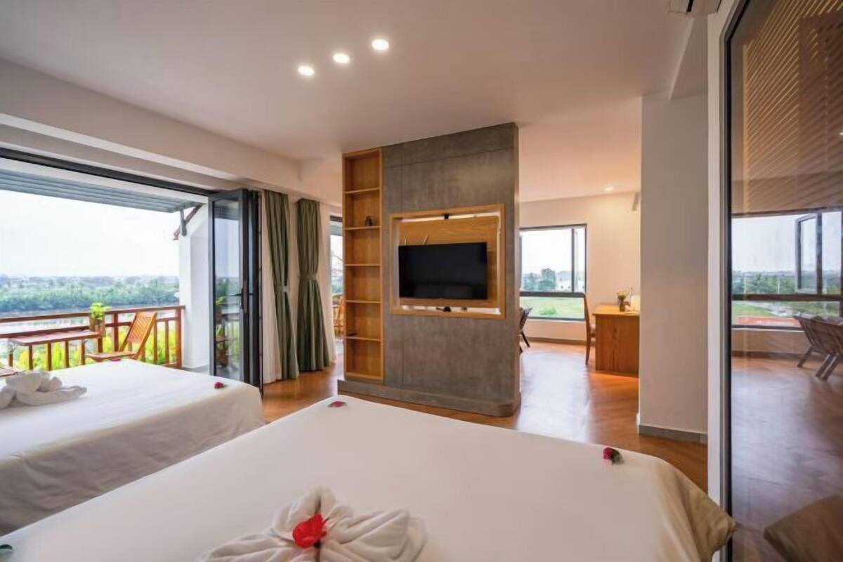 Ein Hotelzimmer mit Balkon, einem Bett mit Blumenarrangement, einem an einer Holzvertäfelung montierten Fernseher und einem Schreibtisch mit Stuhl.