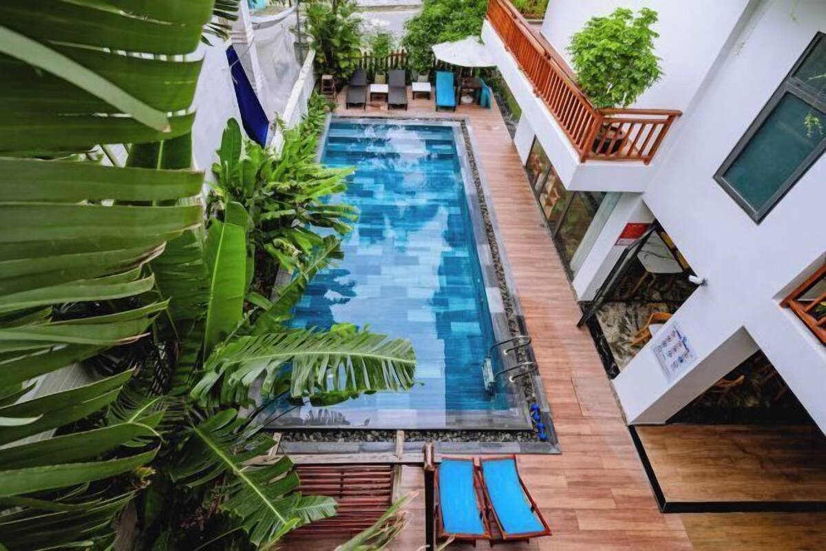Ein rechteckiger Pool mit Holzterrasse, umgeben von üppiger Vegetation und weißen Gebäuden.
