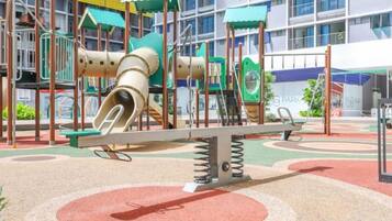 Un terrain de jeux avec un toboggan et des balançoires, situé devant un bâtiment doté d’un auvent bleu.
