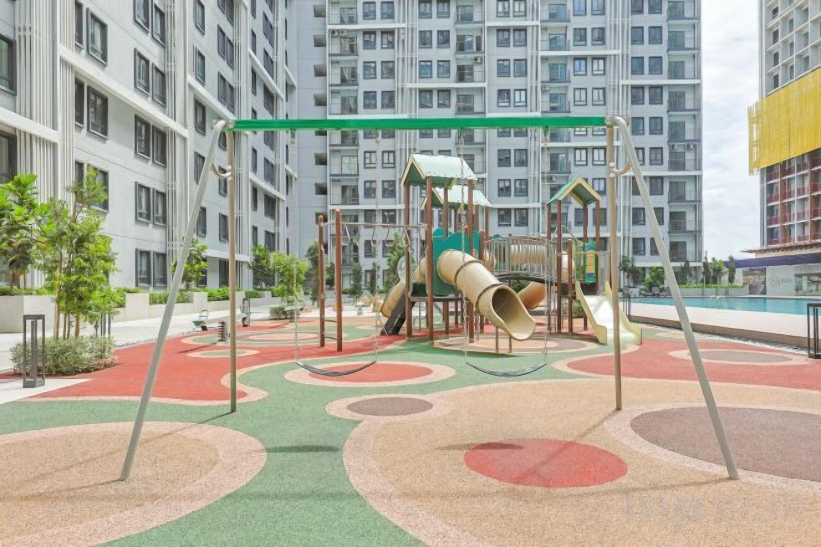 A playground with a slide and climbing structures, surrounded by a green and red play area, with high-rise buildings in the background.