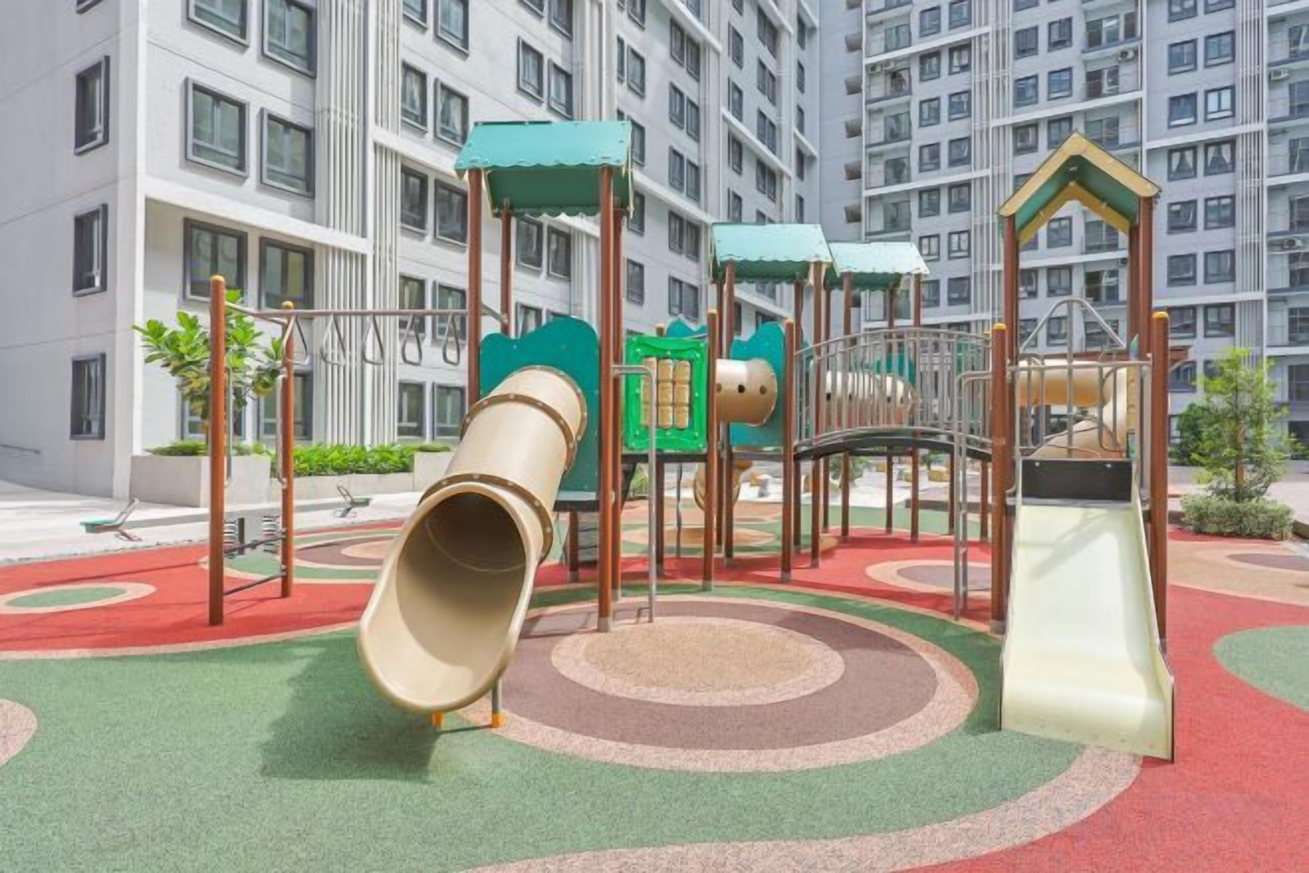 A playground with slides and climbing structures, surrounded by a red and green play area, with a residential building in the background.