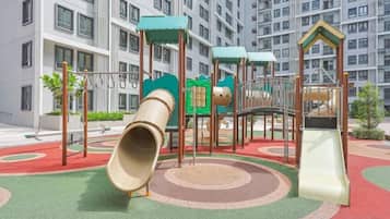 A playground with slides and climbing structures, surrounded by a red and green play area, with a residential building in the background.