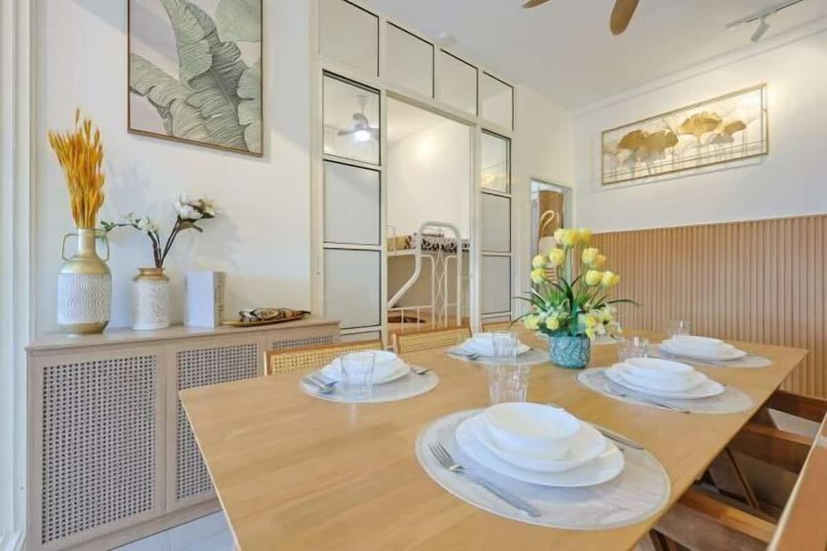Un coin repas avec une longue table en bois dressée pour le repas, bordée de meubles blancs ornés d’objets décoratifs, et un mur où sont accrochées des œuvres d’art encadrées.