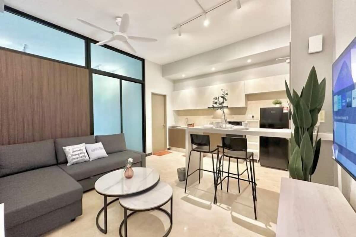 Una sala de estar moderna con sofá, mesa de centro y sillas, una zona de cocina con mesa de comedor y un ventanal grande.