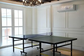 Una mesa de ping pong en una habitación con paneles blancos, un ventanal grande y una araña.