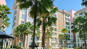 A multi-story residential building with balconies, surrounded by palm trees and a small water feature.