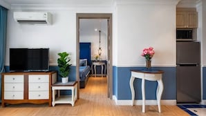 A compact living space with a TV, a small table with a potted plant, a dining area with a lamp, and a kitchen with a refrigerator.