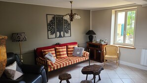 A living room with a red sofa, a black leather couch, a wooden side table, a wicker chair, and a window with a view of greenery.