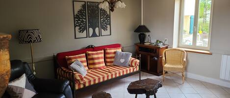 A living room with a red sofa, a black leather couch, a wooden side table, a wicker chair, and a window with a view of greenery.