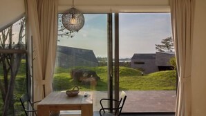 A dining area with a table and chairs, a hanging light fixture, and a view of a modern building and grassy landscape through large glass doors. - Kairosjeju Boutique Stay (Jeju)