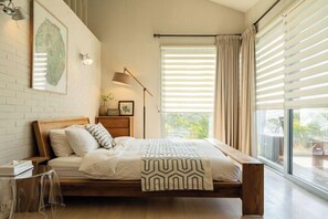 A bedroom with a wooden bed, a bedside table, a lamp, a window with blinds, and a view of greenery outside. - Kairosjeju Boutique Stay (Jeju)