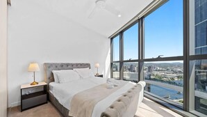 Une chambre à coucher moderne, dotée d’un grand lit, de deux tables de chevet et offrant une vue sur le paysage urbain grâce à de grandes fenêtres.