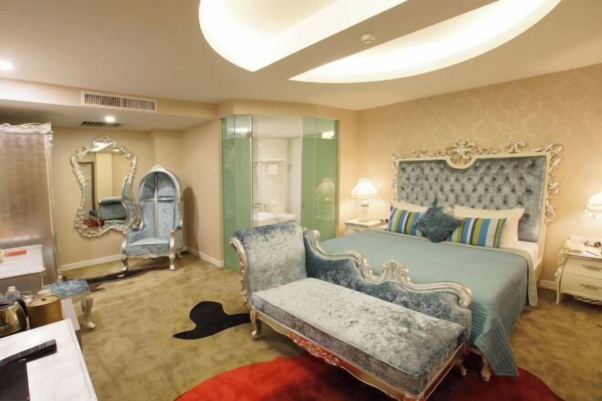 Un dormitorio con una cama grande, un tocador con espejo, un sillón azul y una alfombra roja.