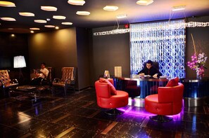 Un espace d’accueil d’hôtel comprenant un comptoir de réception, deux fauteuils rouges, un grand mur décoratif et un sol à motifs foncés.