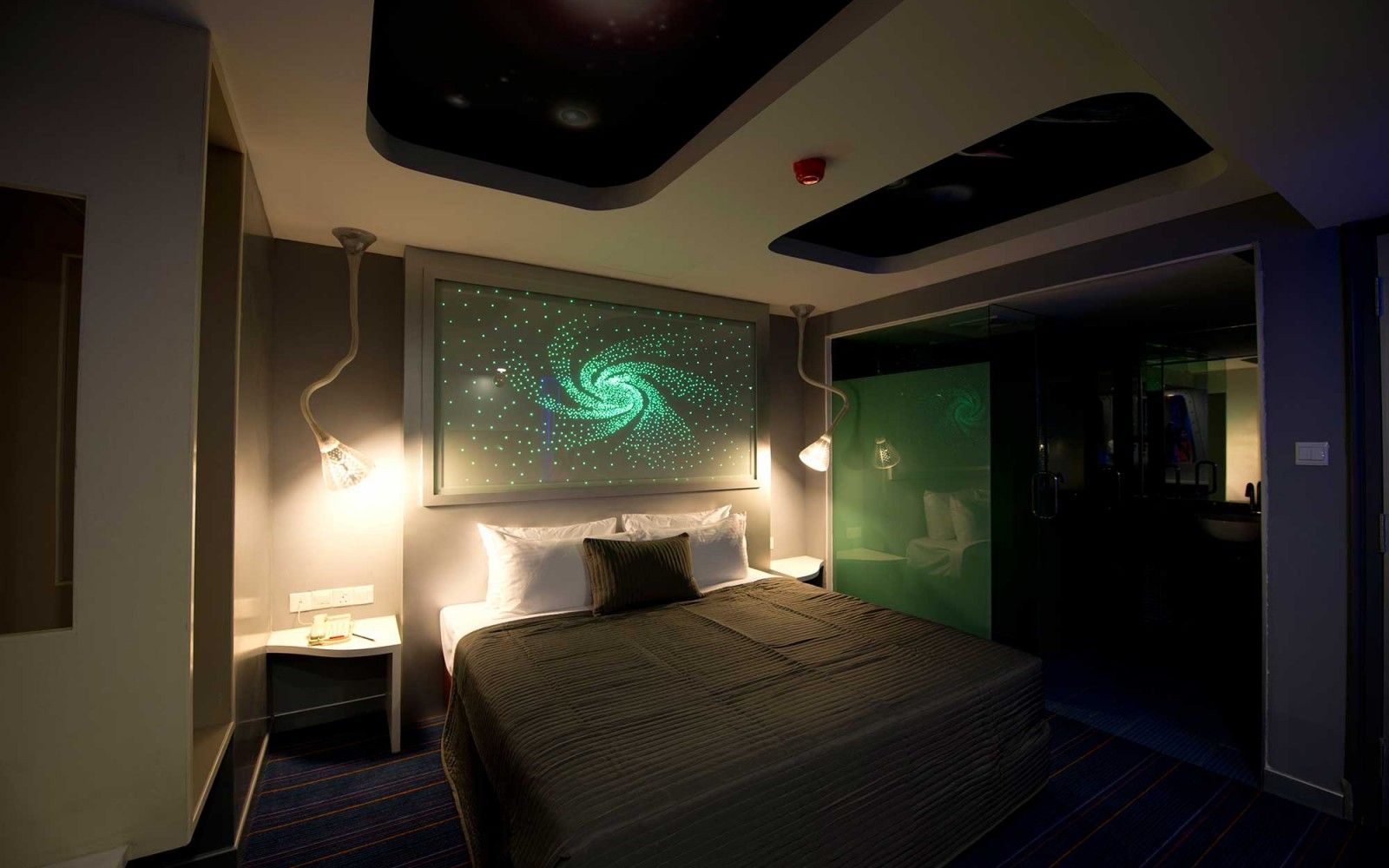 A hotel room with a bed, a wall-mounted projector displaying a green spiral, and a nightstand with a lamp.