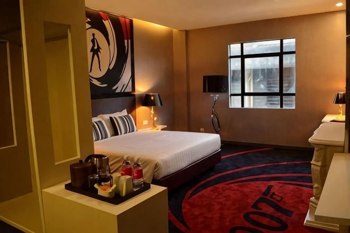 Une chambre d’hôtel avec un lit, une table de chevet, une lampe, une fenêtre et un tapis rouge orné d’un logo.