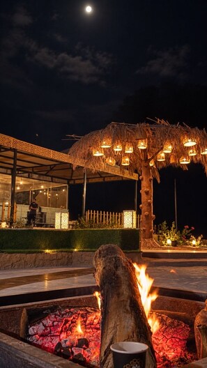 A nighttime outdoor area with a fire feature, a thatched structure with lights, and a building with a glass facade.