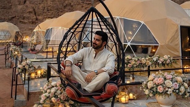 Un homme est assis dans un fauteuil suspendu, avec en arrière-plan un paysage désertique, des tentes et des compositions florales.