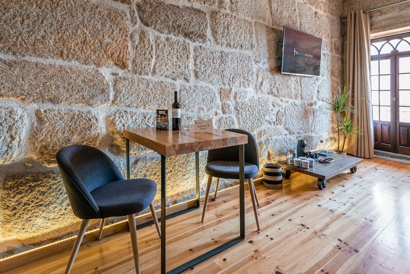 A rustic room with a stone wall, a wooden table with two chairs, a television mounted on the wall, and a window with curtains.