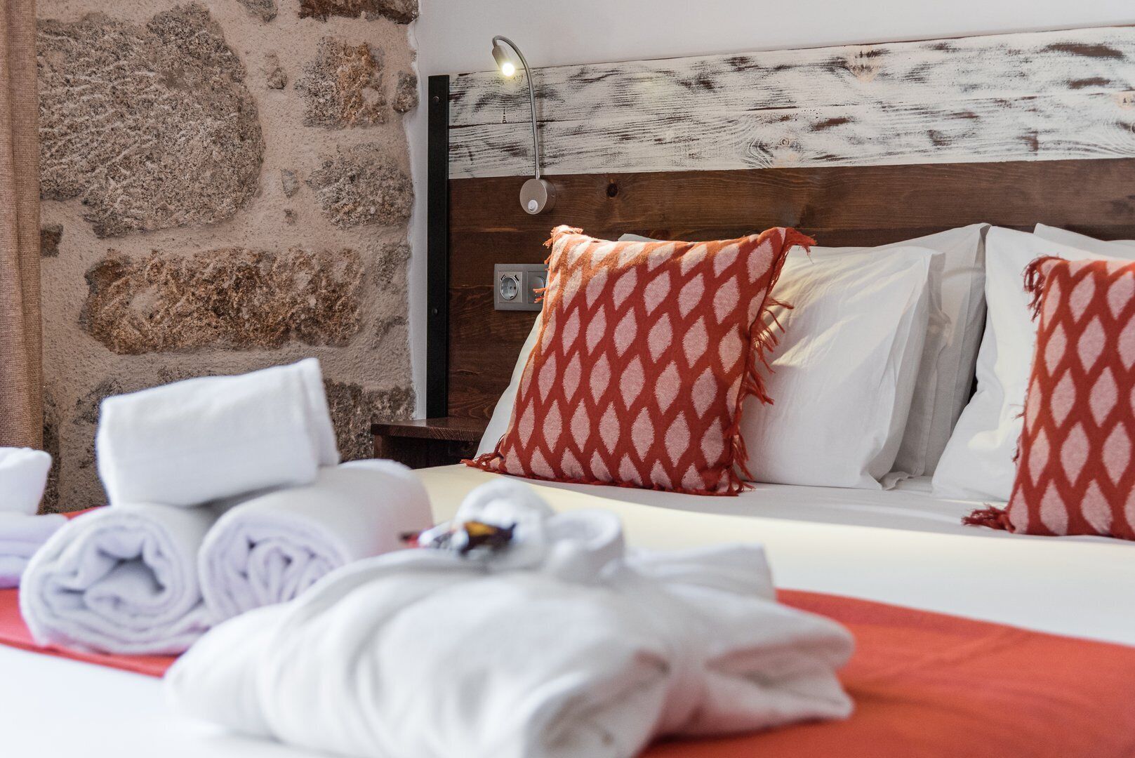 A hotel room with a stone wall, a wooden headboard, and a bed with white and red patterned pillows. There are towels and a remote control on the bed.