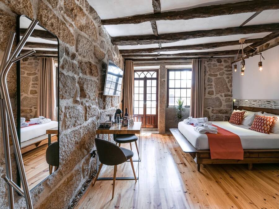 Una habitación rústica con paredes de piedra vista, piso de madera, cama con una manta roja, una mesa de comedor con sillas y un televisor montado en la pared.