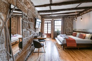 A rustic room with exposed stone walls, a wooden floor, a bed with a red blanket, a dining table with chairs, and a TV mounted on the wall. - Guestready - Castlerock Pub Hotel (Porto)