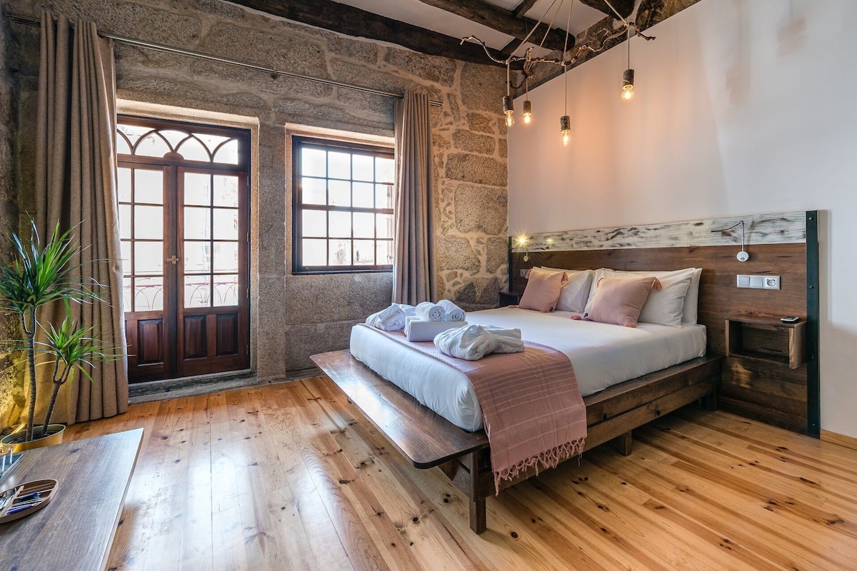 A bedroom with a stone wall, wooden floor, a bed with pillows and towels, a bench, and a bedside table.