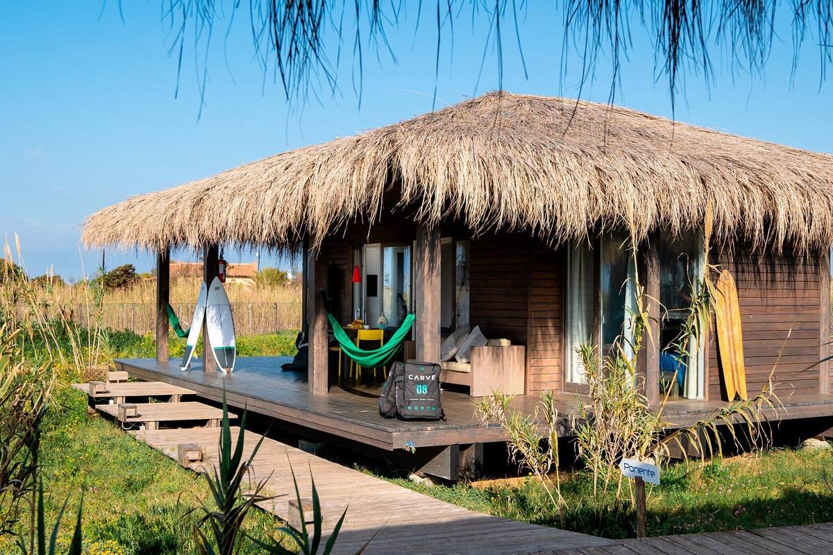 A thatched-roof bungalow with a wooden deck, a hammock, and a surfboard leaning against the wall.