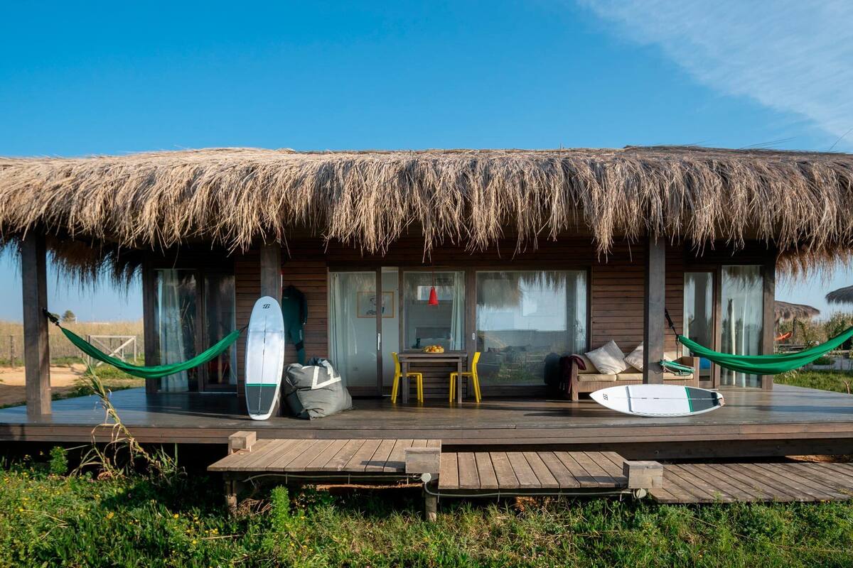A thatched-roof building with a wooden deck, two hammocks, and surfboards.