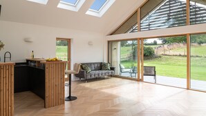 Un vaste espace de vie agrémenté d’un grand parquet à motifs en chevrons, d’un canapé gris, d’une table à manger en bois sur laquelle sont disposés des fruits, et offrant une vue sur un paysage verdoyant grâce à de grandes fenêtres.