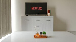 A modern room with a white cabinet, a television displaying 'NETFLIX,' and a table with a tray holding a plant and candles. - Calma 2BR II @Skyhouse Apartment (Cisauk)
