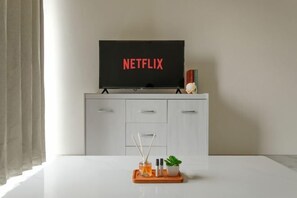 A modern room with a white cabinet, a television displaying 'NETFLIX,' and a table with a tray holding a plant and candles. - Calma 2BR II @Skyhouse Apartment (Cisauk)