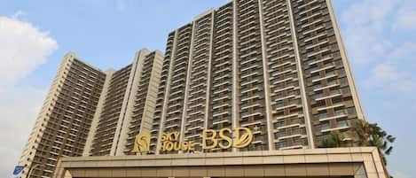 Un edificio residencial de gran altura con múltiples balcones, un cartel llamativo que dice 'Sky House BSD' y una entrada visible con un letrero de 'Rishi AID'.