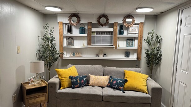 A living room with a grey sofa, yellow and patterned cushions, wooden side tables, and decorative items on shelves. - Convenient 1-BR - Fenced Backyard - Extended Stays wanted (Huntington)