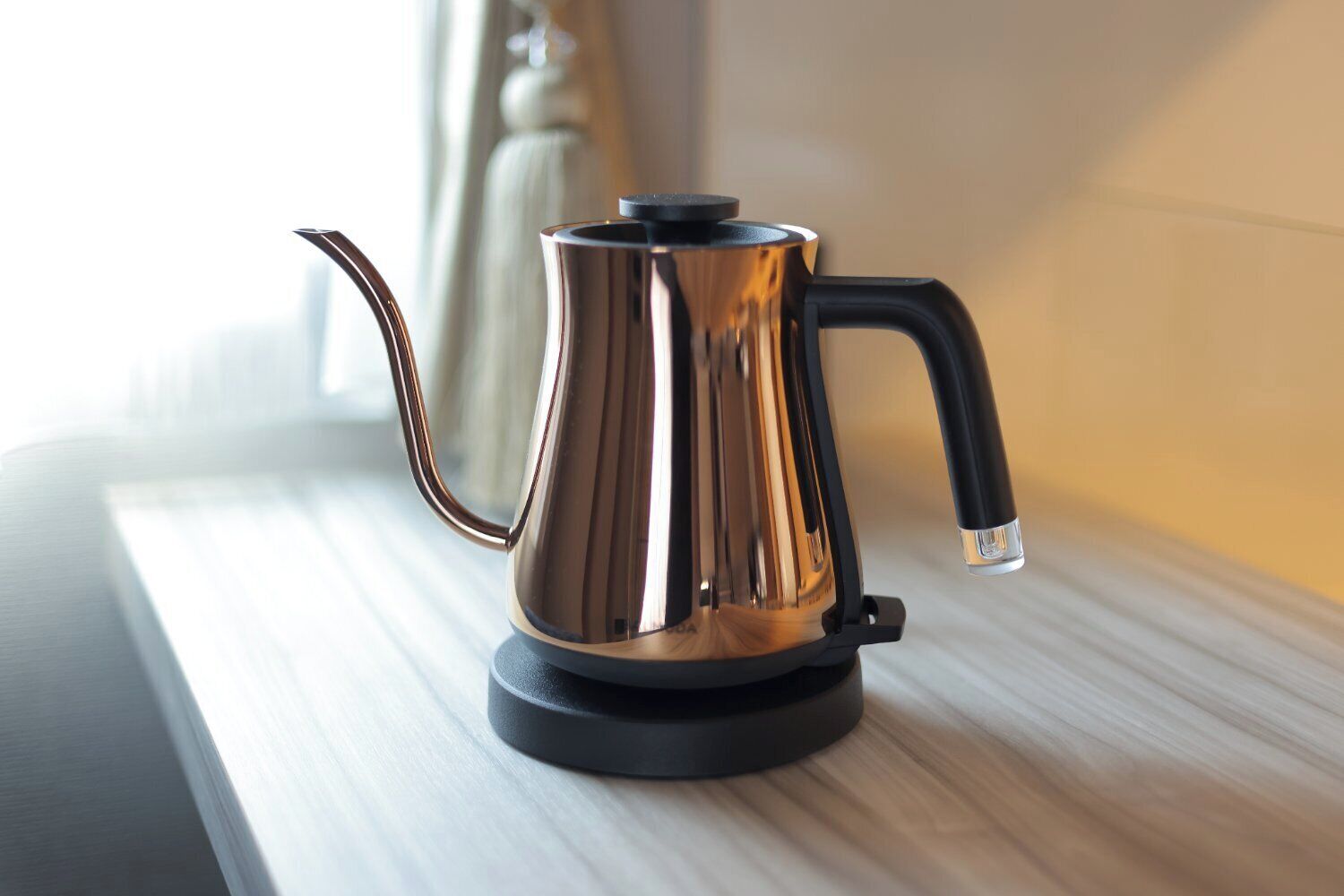 A stainless steel kettle on a black base, placed on a wooden surface near a window.