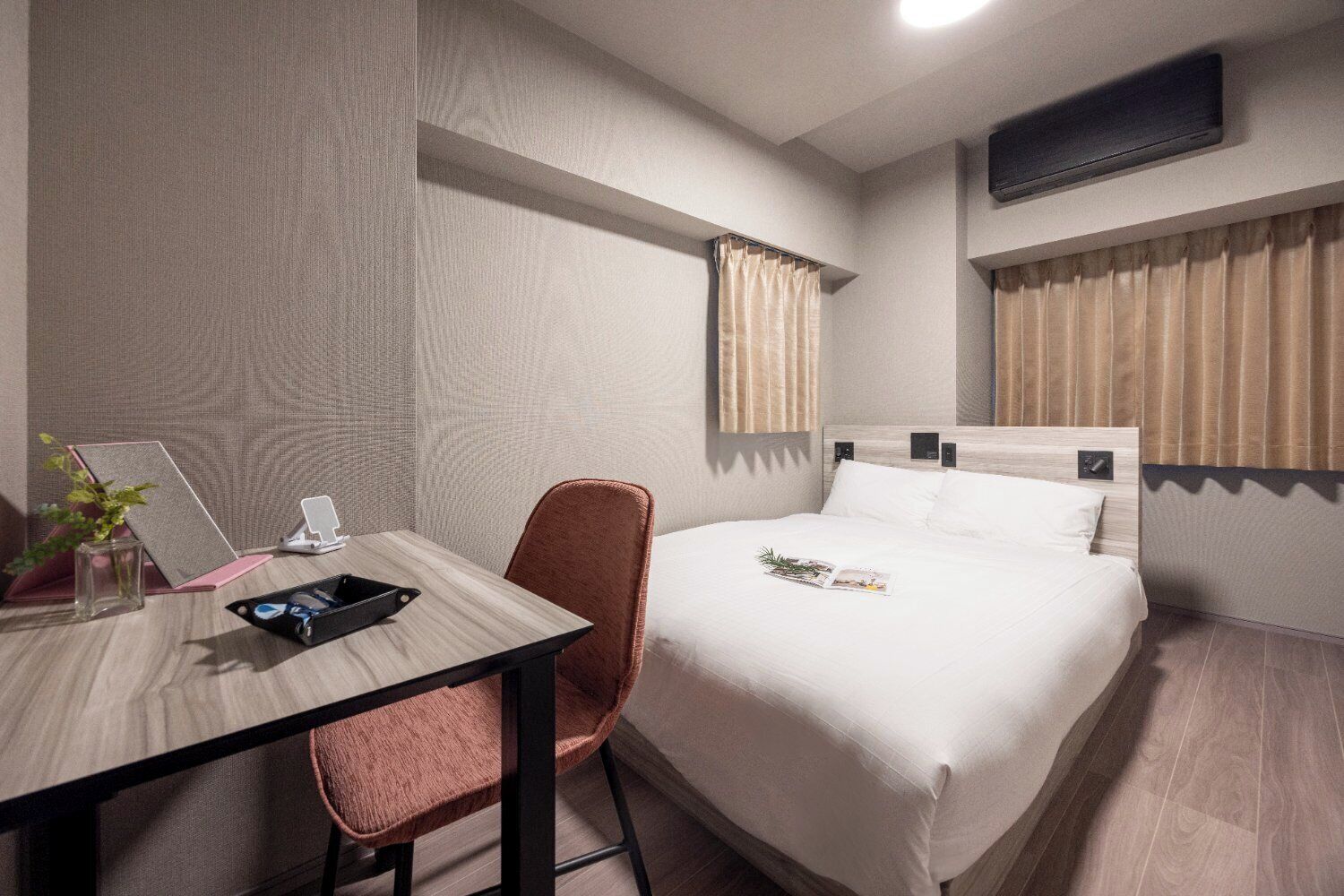 A hotel room with a bed, a desk with a chair, a small table with a plant, and a window with curtains.