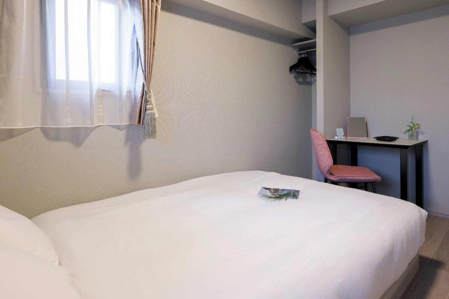 A neatly made bed with a magazine on it, a chair, a desk with a laptop, and a window with sheer curtains.