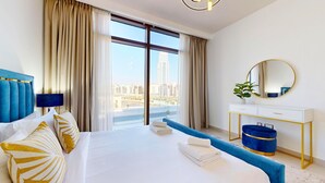 A hotel room with a large bed, a blue headboard, a white dresser with a mirror, and a view of a cityscape through large windows. - Cove Residences - Creek Harbour Dubai (Dubai)