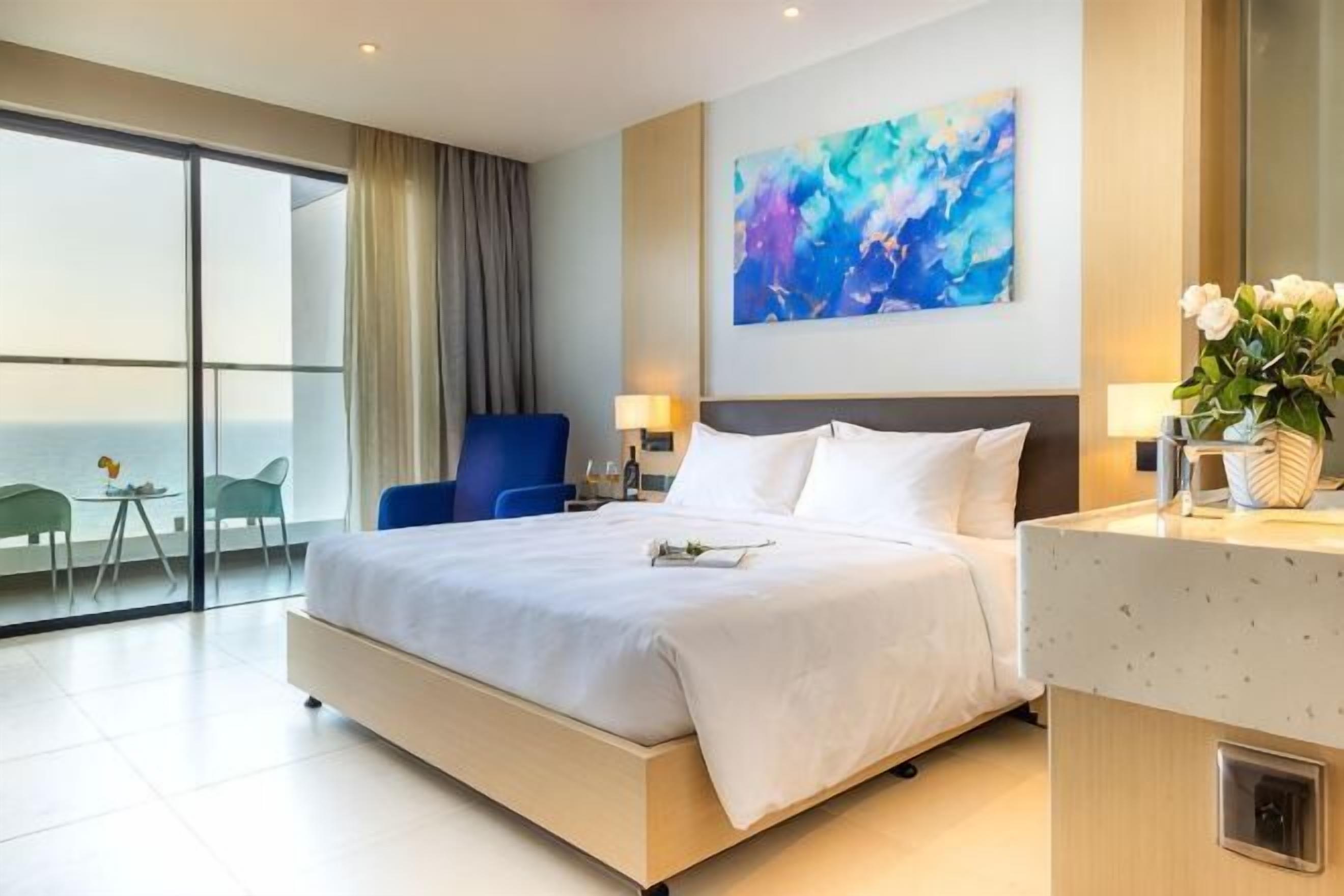Ein Hotelzimmer mit einem großen Bett, einem Schreibtisch, einem Sessel und Meerblick. An der Wand hängt ein Gemälde.