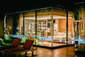 Una habitación moderna con paredes de vidrio, piscina, sillas de descanso y una cama.