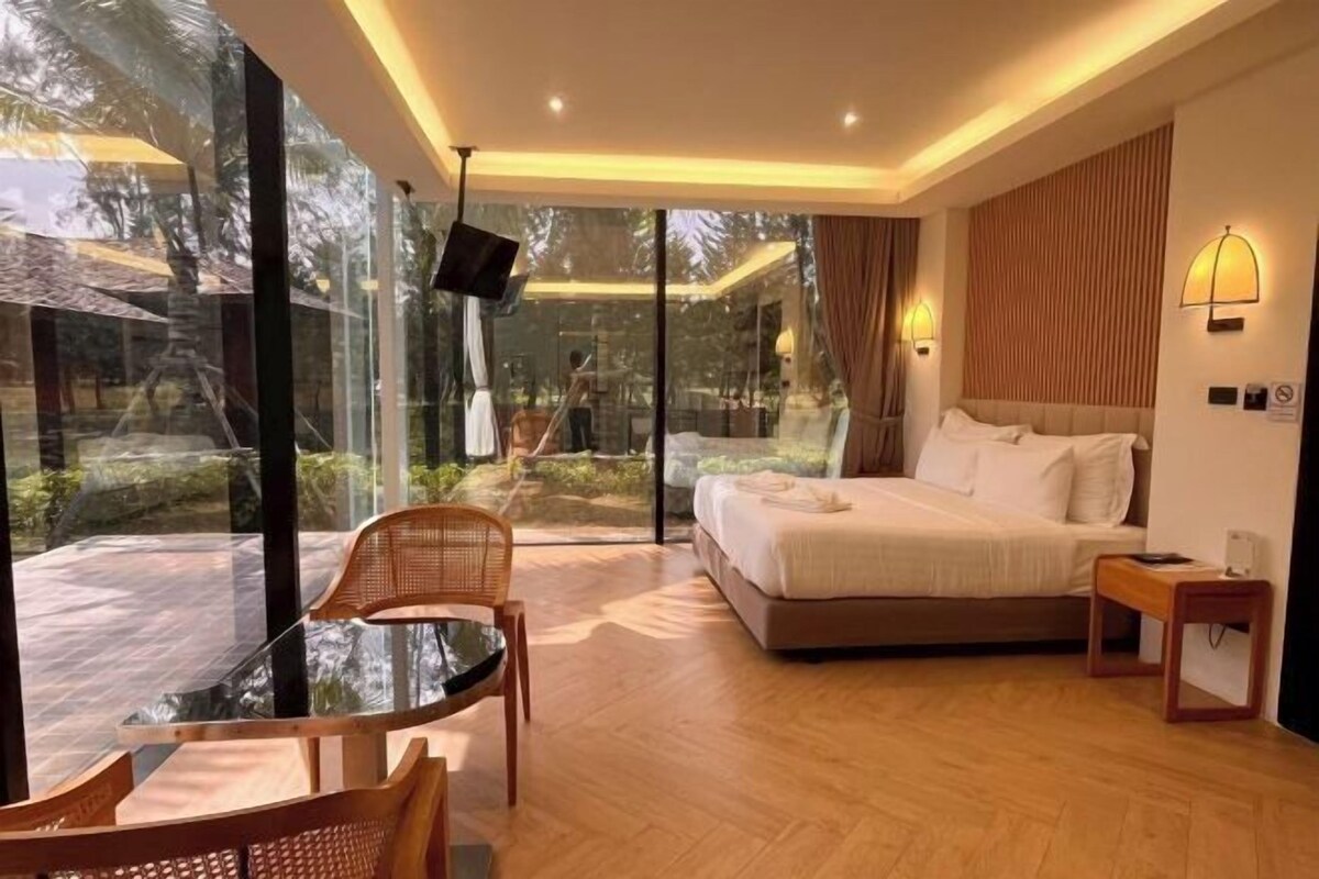 A modern hotel room with a large bed, a sitting area with a glass table, and a view of the outdoors.