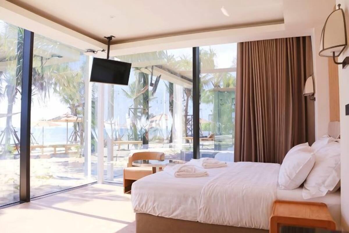 A bedroom with a bed, a TV, a window, and a view of the beach.