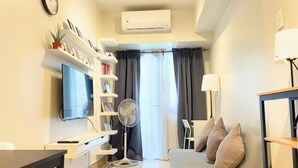 A modern living room with a flat-screen TV, a standing fan, a sofa with cushions, a side table, and a lamp. - Callie's Cozy Staycation (Pasay)