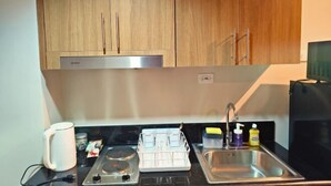 A compact kitchen with wooden cabinets, a stainless steel sink, a kettle, a dish rack, and a coffee maker. - Callie's Cozy Staycation (Pasay)